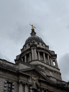 Upward-looking photo of the Old Bailey’s dome in London, featuring the golden Lady Justice statue holding a sword and scales against an overcast sky.
