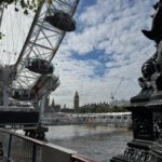 A view of the London Eye and Big Ben from the South Bank, with the Thames, riverside railing, and decorative lamp sculpture in the foreground.