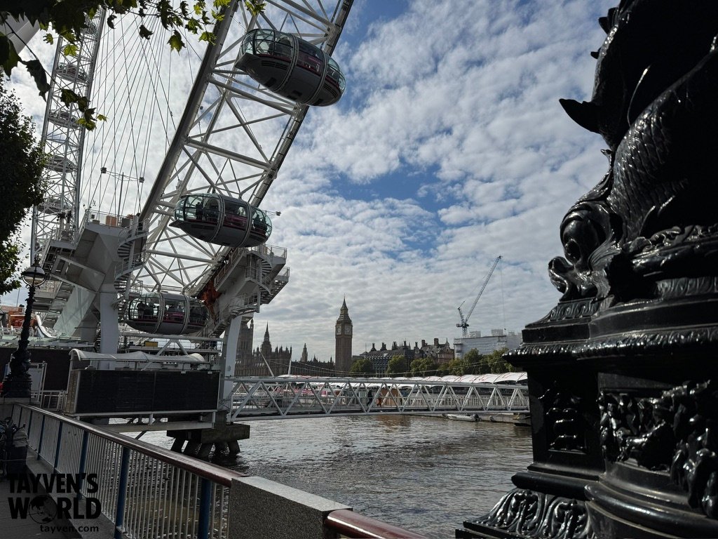 A view of the London Eye and Big Ben from the South Bank, with the Thames, riverside railing, and decorative lamp sculpture in the foreground.