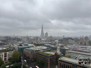 Wide cityscape photo of London showing The Shard rising above surrounding buildings, with the River Thames and bridges visible under an overcast sky.