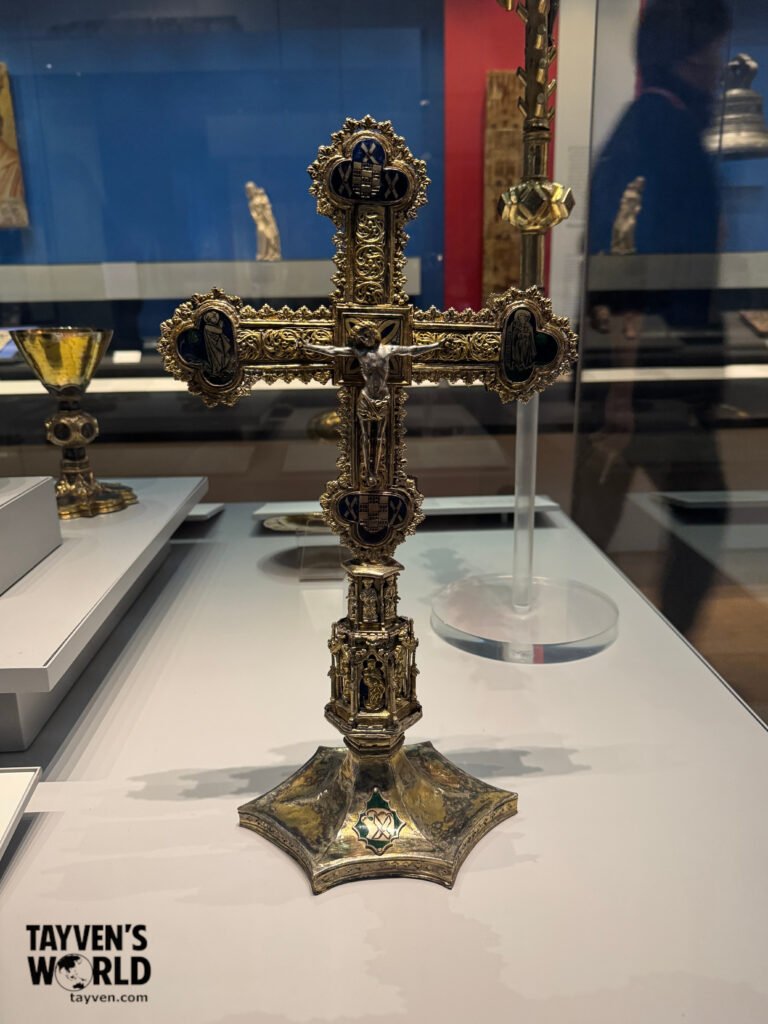 Gilded Medieval Cross Ornate gilded cross with enamelled religious figures displayed in a museum case.