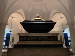 Black sarcophagus with gold detailing marking the tomb of Admiral Horatio Nelson, set beneath vaulted white arches with hanging lamps in the crypt of St Paul’s Cathedral.