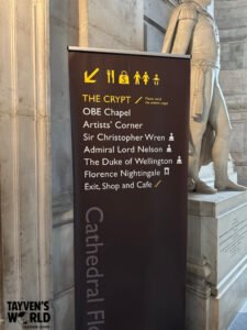 Photo of an interior cathedral sign pointing toward the crypt, listing areas such as the OBE Chapel, Artists’ Corner, and tombs of notable figures, with part of a statue visible behind it.