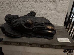 Carved effigy of William Hewit lying atop a stone memorial base, with inscriptions noting his death in 1599, set against a textured cathedral wall.
