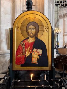 Icon of Jesus Christ in a cathedral, with a gold background, halo markings, and lit candles below, framed by tall stone columns.