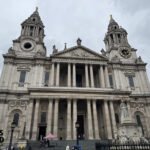 Wide-angle photo of the front entryway of St Paul’s Cathedral in London, showing the stone steps leading up to the large wooden doors and the symmetrical twin towers above.