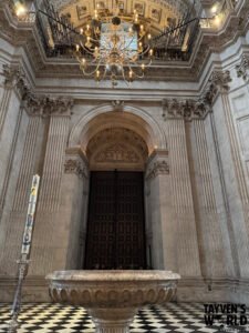 Interior photo of a cathedral entrance showing a marble baptismal font, tall columns, a large wooden door, a Paschal candle, and an ornate chandelier above.