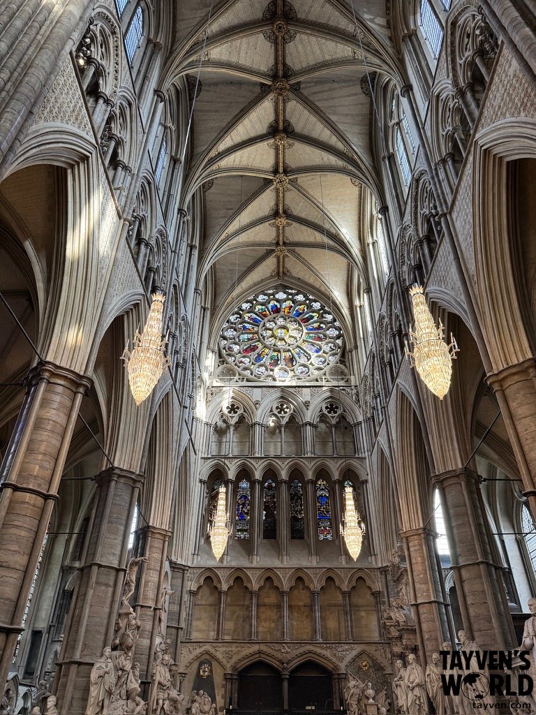 Stained‑glass rose window and vaulted Gothic ceiling inside Westminster Abbey, with chandeliers and carved stone figures along the walls.