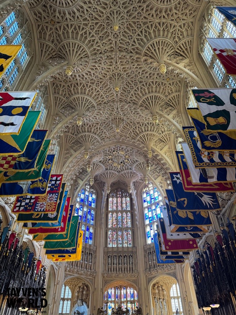 Interior of St George’s Chapel at Windsor Castle with fan‑vaulted ceiling, stained‑glass windows, and colourful Garter banners hanging above the nave.