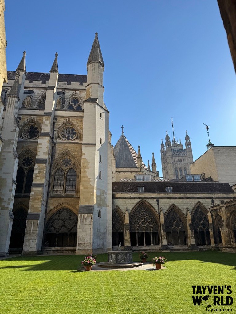 Cloister courtyard at Westminster Abbey with Gothic arches, stone tracery, and a small central fountain under a clear blue sky.