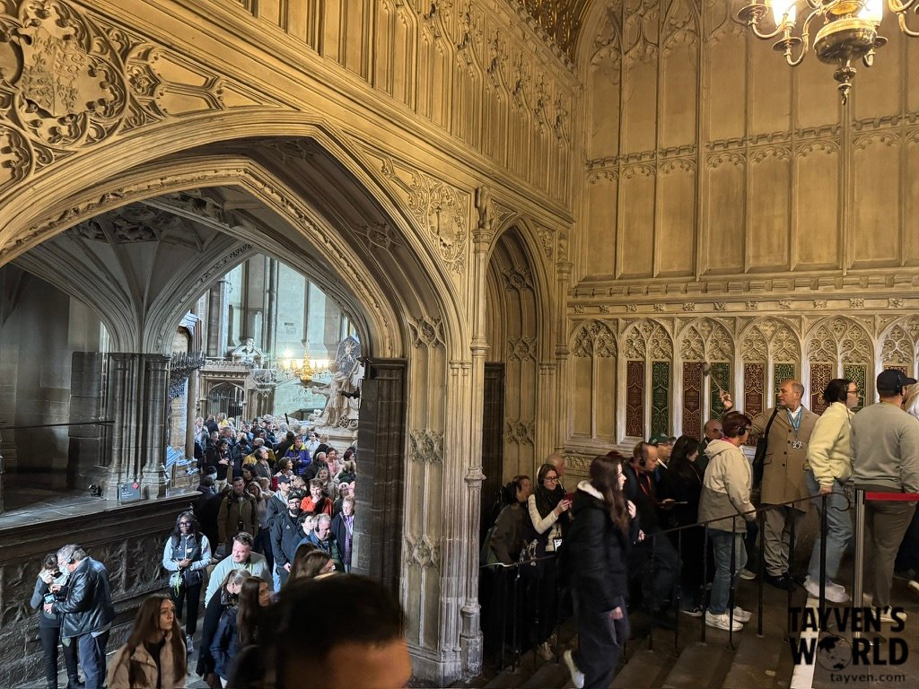 Crowds of visitors inside Westminster Abbey, surrounded by ornate Gothic stonework, pointed arches, and illuminated chandeliers.