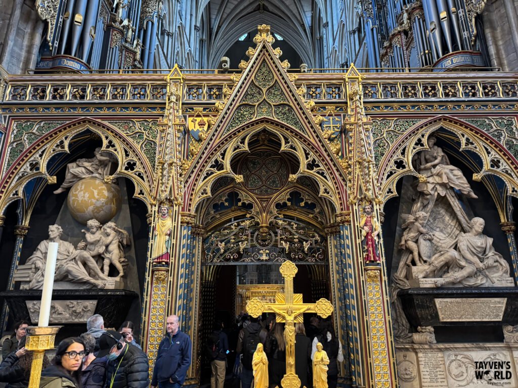 Golden cross and surrounding stone sculptures inside Westminster Abbey, with organ pipes and Gothic arches in the background.