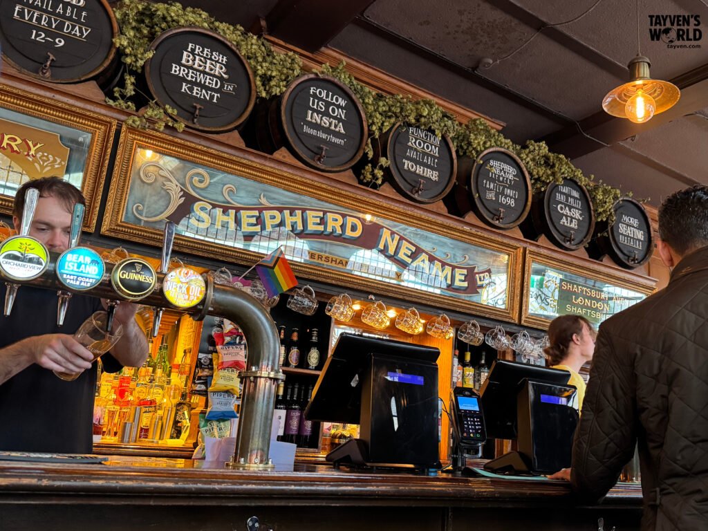Bartender pouring beer at The Bloomsbury Tavern in London, with vintage Shepherd Neame signage and wooden barrels above the bar.