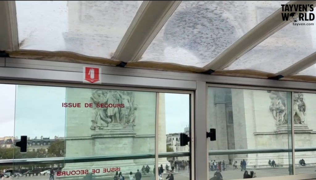 View of the Arc de Triomphe in Paris seen through the glass window of a covered tour bus, with “Issue de Secours” emergency‑exit text reflected on the glass.