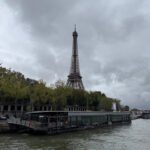 Eiffel Tower viewed from across the Seine River with boats and trees in the foreground.