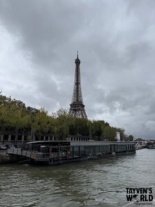 Eiffel Tower viewed from across the Seine River with boats and trees in the foreground.