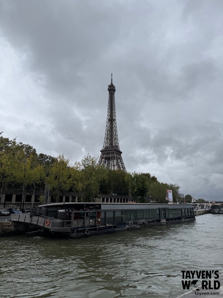 Eiffel Tower viewed from across the Seine River with boats and trees in the foreground.