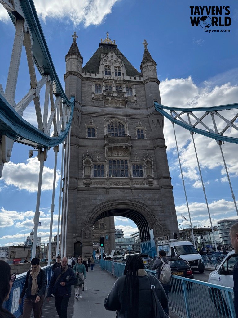 Pedestrians crossing Tower Bridge with its Gothic towers and blue steel framework.