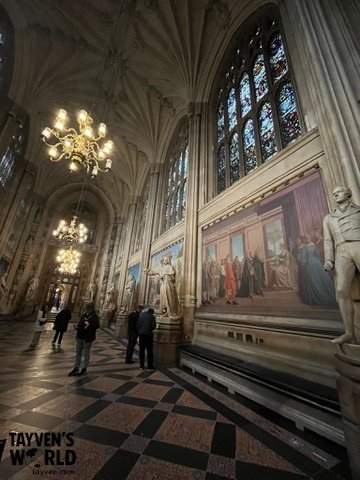 Long Gothic corridor with stained glass windows, murals, and statues inside Westminster Palace.