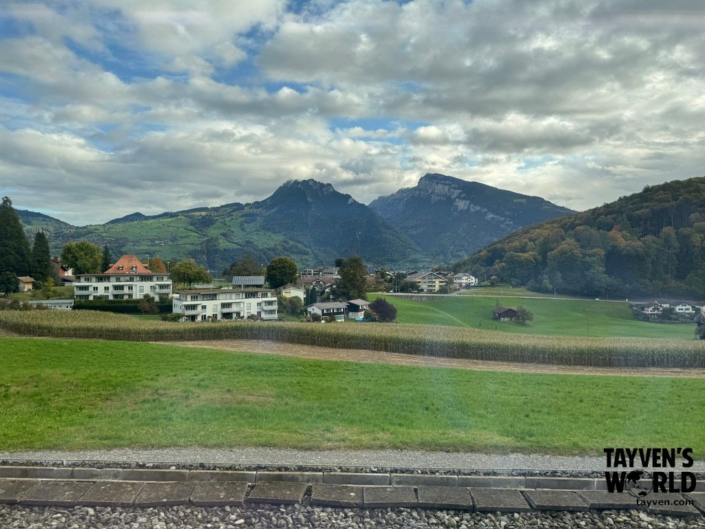 Green fields and houses with mountains rising behind under a partly cloudy sky.