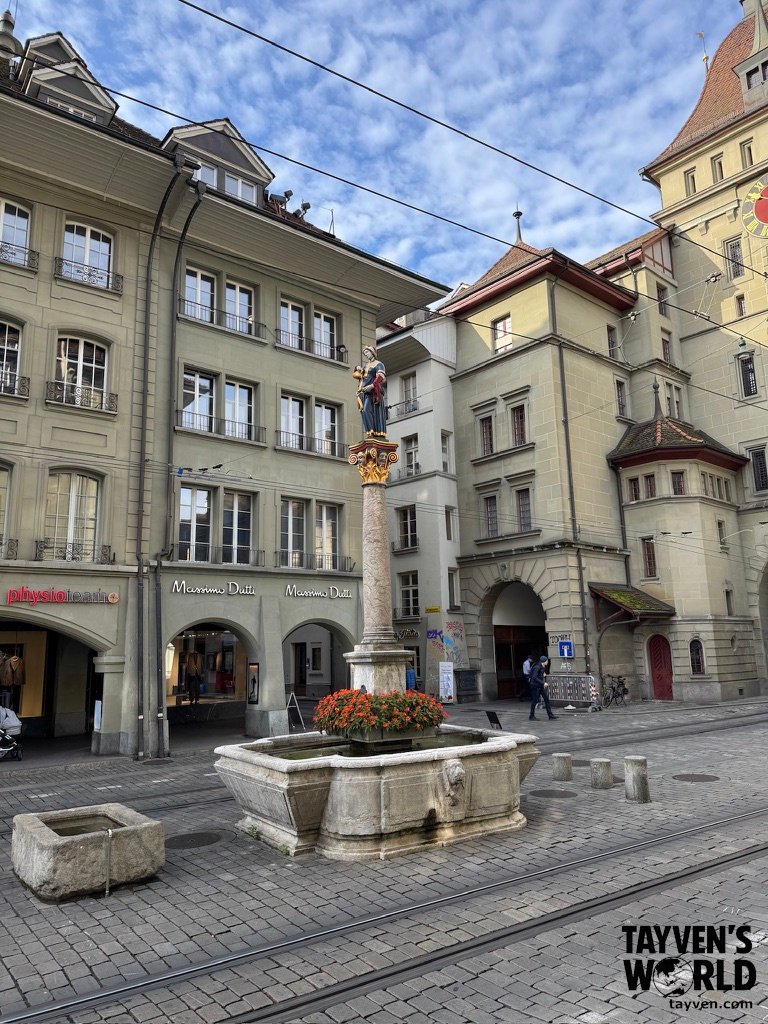 Stone fountain with floral base and statue in Bern’s old town, surrounded by shops and tram tracks.