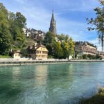Riverside view of Bern with turquoise water, hillside homes, and the tall cathedral spire rising above.