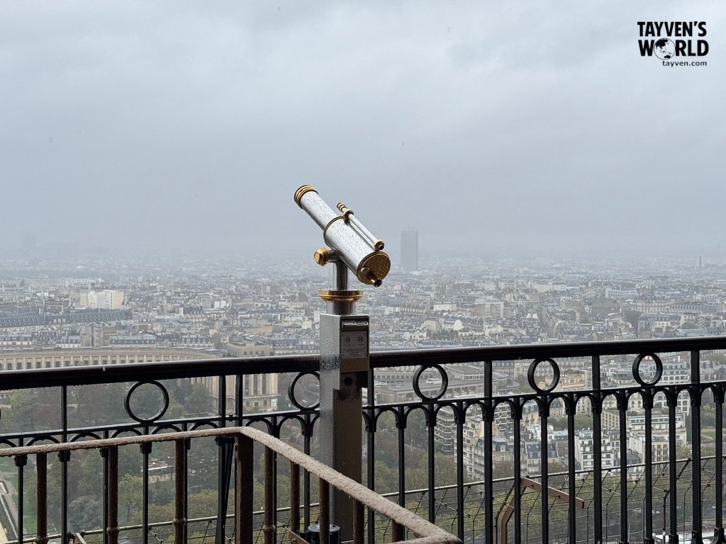 Telescope overlooking Paris from the Eiffel Tower observation deck on a cloudy day.