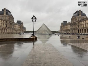 Louvre courtyard with glass pyramid and wet cobblestones on a rainy day.