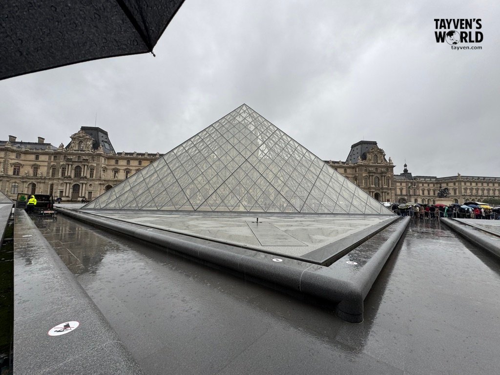 Close‑up view of the Louvre glass pyramid on a rainy day with umbrellas in the crowd.
