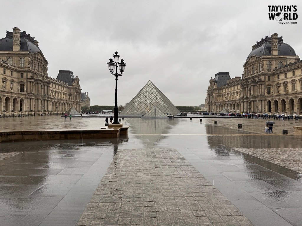 Louvre courtyard with glass pyramid and wet cobblestones on a rainy day.