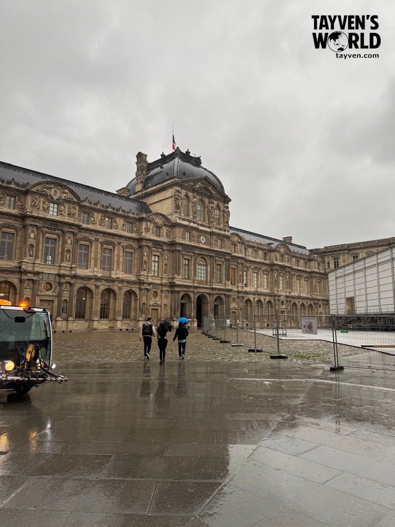 Louvre Museum exterior with wet cobblestones and visitors walking under umbrellas.