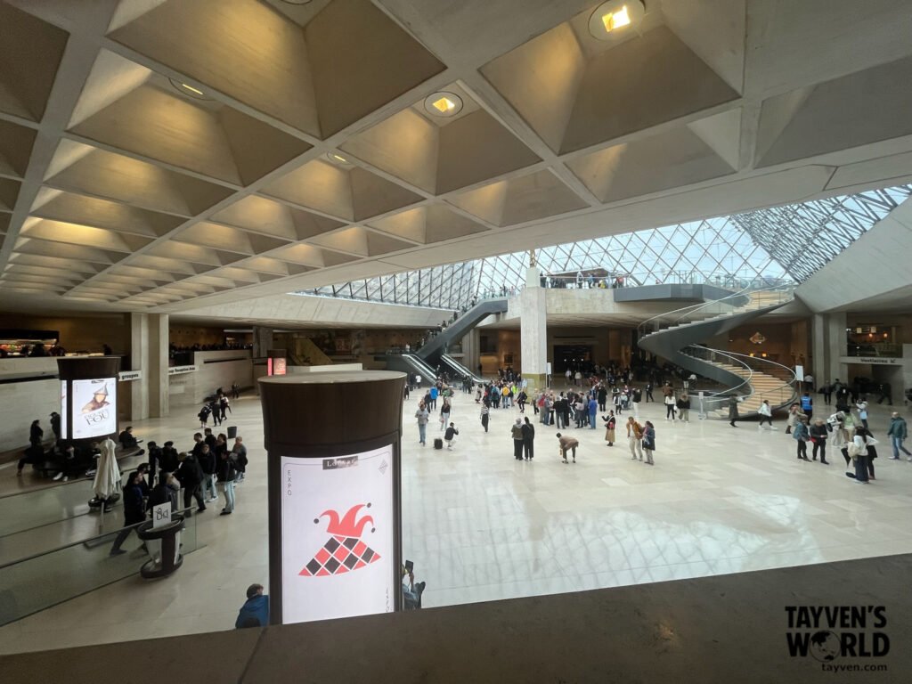 Interior view of the Louvre Pyramid entrance with visitors and spiral staircase.