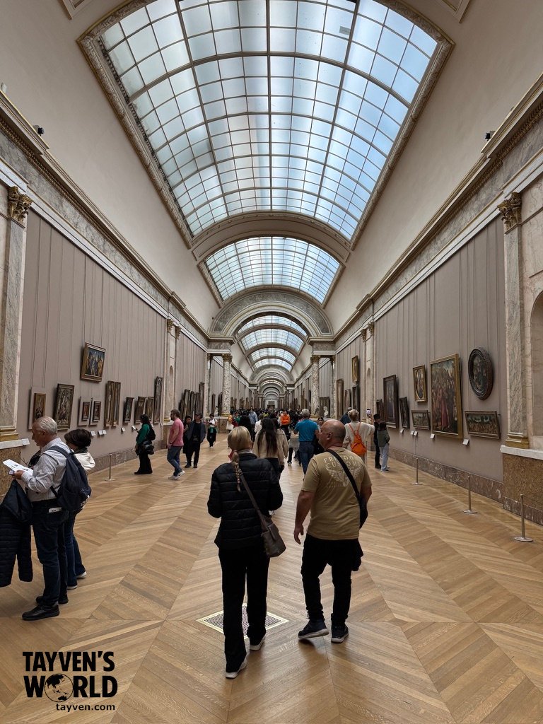 Visitors walking through the Louvre’s long gallery hall lined with paintings.