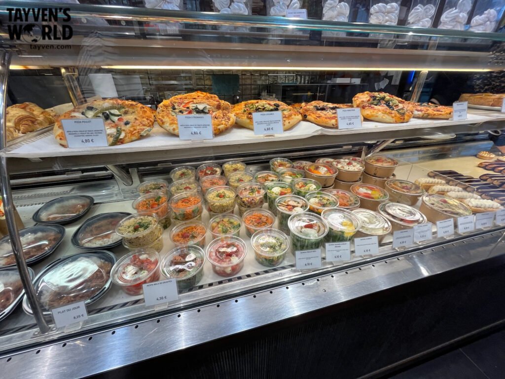 Bakery display with pizzas, salads, and prepared meals labeled with prices in euros.
