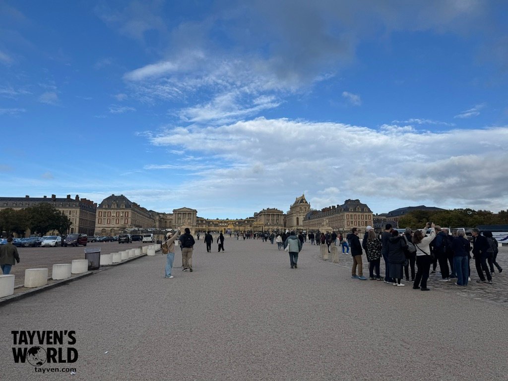 Wide view of visitors approaching the Palace of Versailles under a cloudy sky.