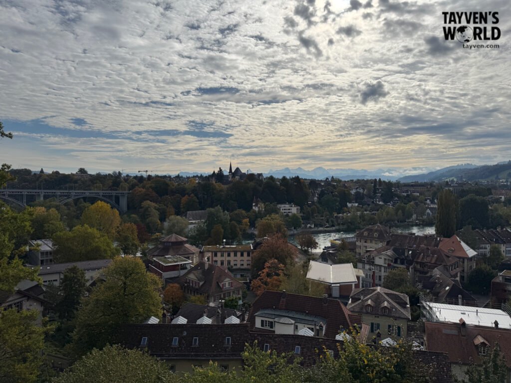 Panoramic view of Bern with the Aare River, autumn trees, and distant Alps under a cloudy sky