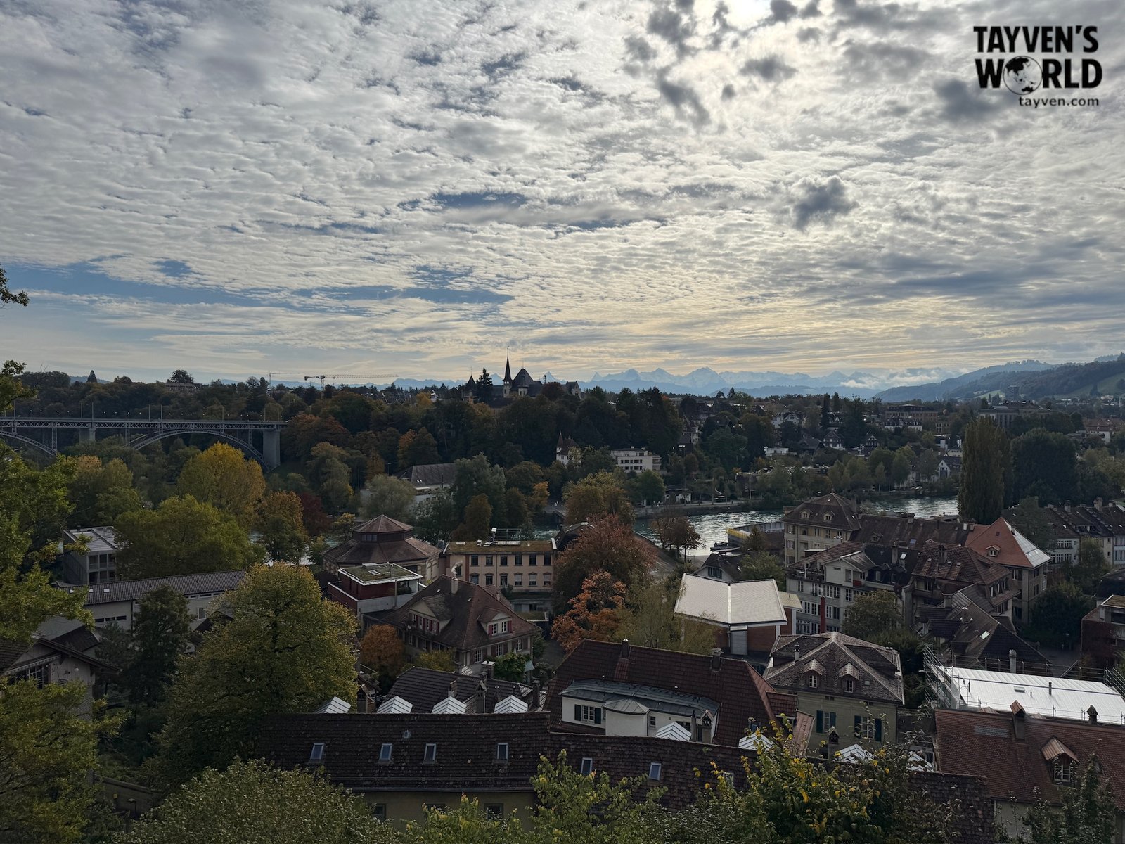 Panoramic view of Bern with the Aare River, autumn trees, and distant Alps under a cloudy sky