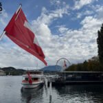 Swiss flag and docked boat on Lake Lucerne with Ferris wheel and mountains in the background.