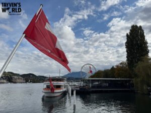Swiss flag and docked boat on Lake Lucerne with Ferris wheel and mountains in the background.