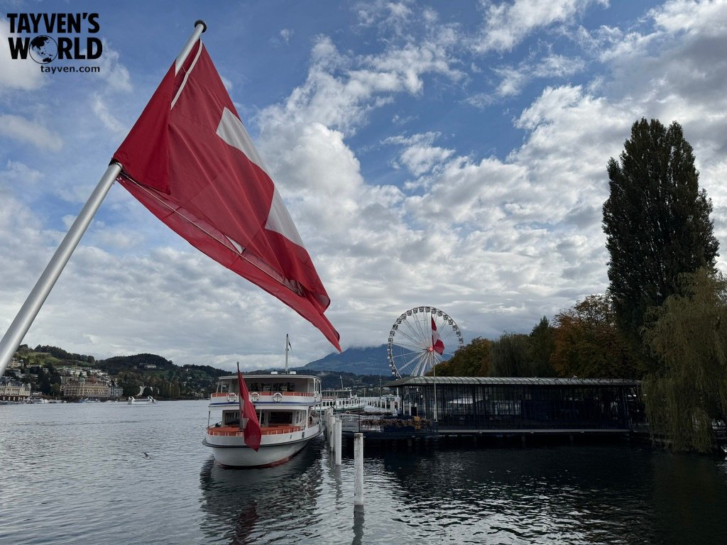 Swiss flag and docked boat on Lake Lucerne with Ferris wheel and mountains in the background.