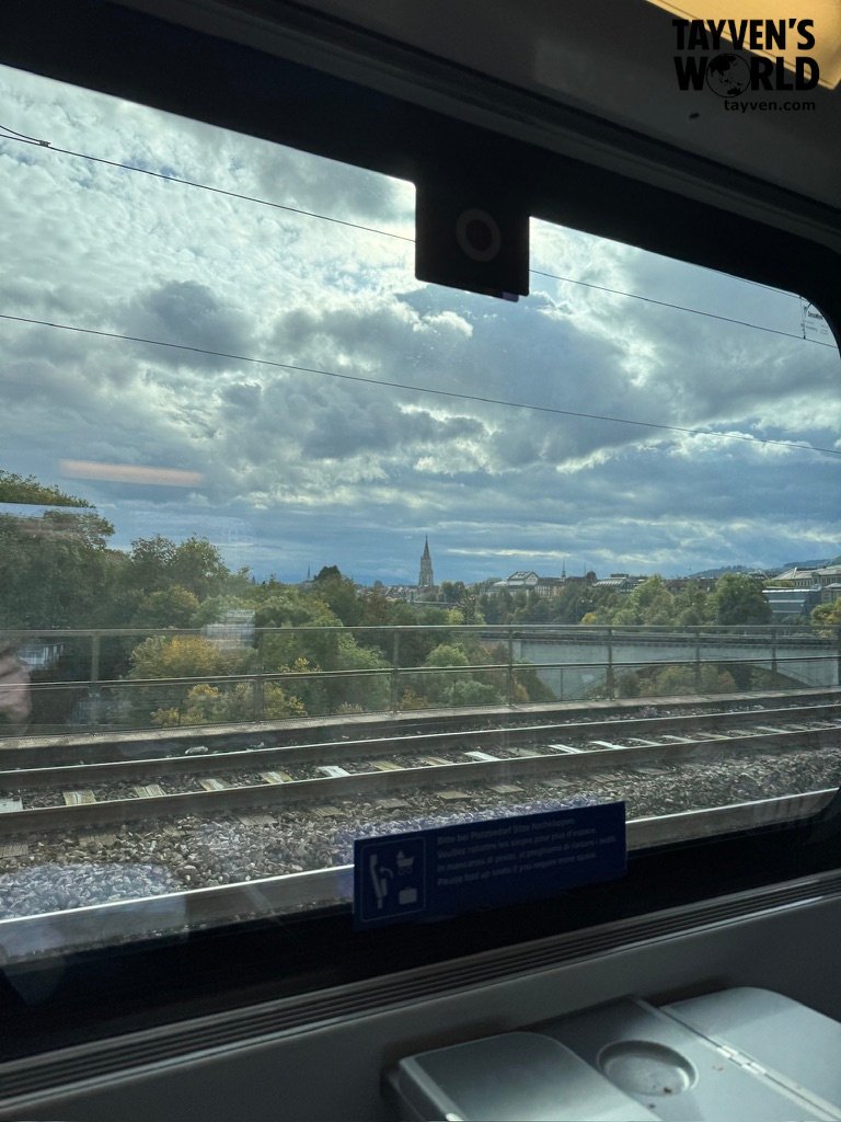 View from a Swiss train window showing railway tracks, autumn trees, and a distant church steeple under dramatic clouds.