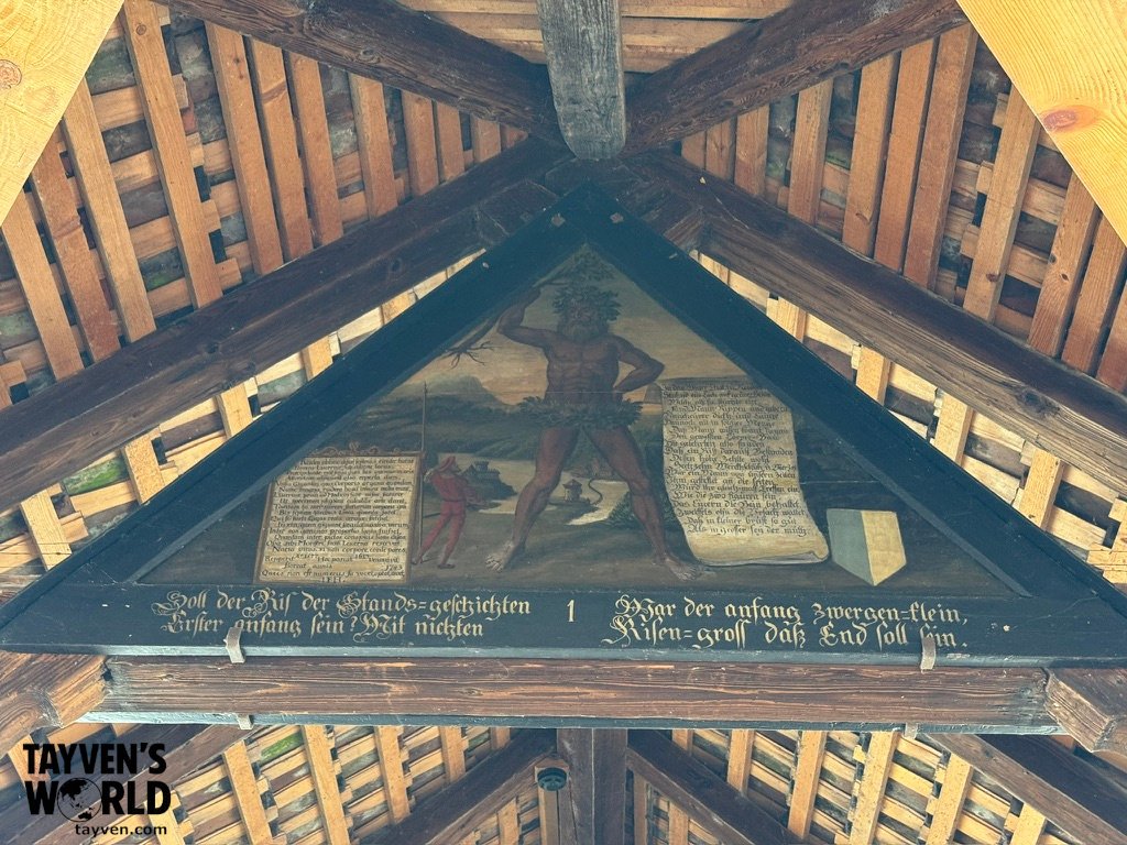 Historic triangular wooden panel with German text and a painting of a giant and a smaller figure under a timber roof.