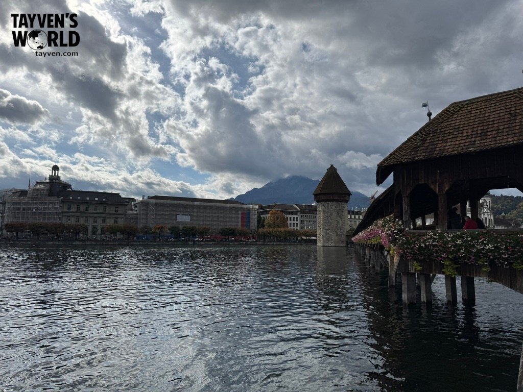 Lucerne’s Chapel Bridge and Water Tower over calm water with flowers and mountains in the background.