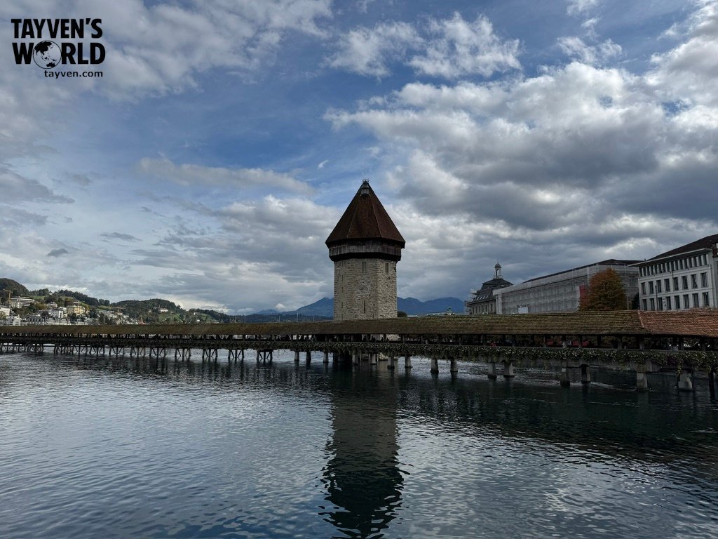Wide view of Lucerne’s Chapel Bridge and Water Tower reflected in the Reuss River under a cloudy sky.