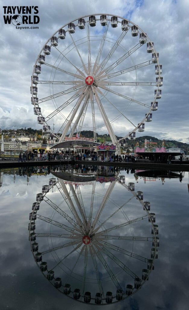 Large Ferris wheel with Swiss cross hub reflected perfectly in calm water at Lucerne’s lakeside.