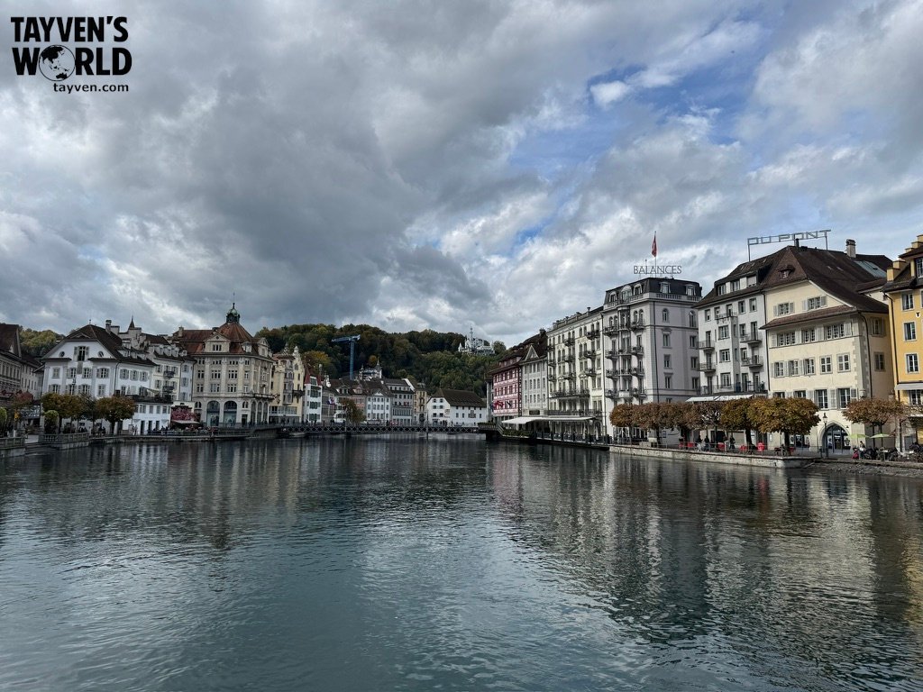 Riverside view of Lucerne’s historic buildings reflected in the Reuss River under a cloudy sky.
