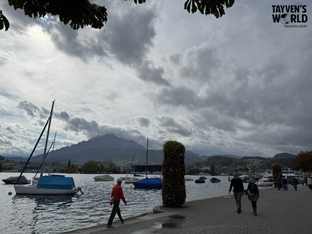 Lakeside promenade in Lucerne with sailboats, people walking, and mountains under a cloudy sky.