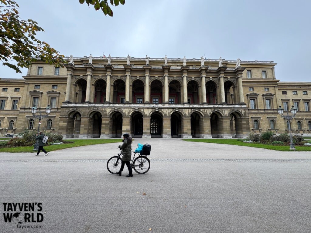 Historic arched building beside Munich’s Hofgarten with a cyclist passing in front Title: Historic Architecture Near Munich’s Hofgarten