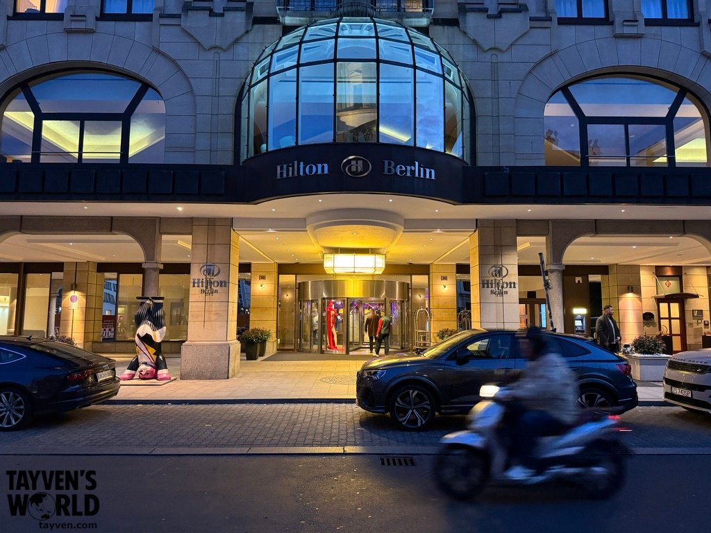 Illuminated entrance of the Hilton Berlin hotel with vehicles passing outside at dusk